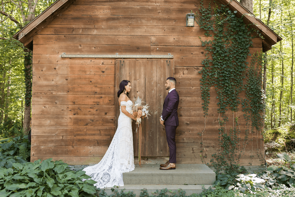 Bride and groom posing in front of wooden barn while bride holds her bouquet